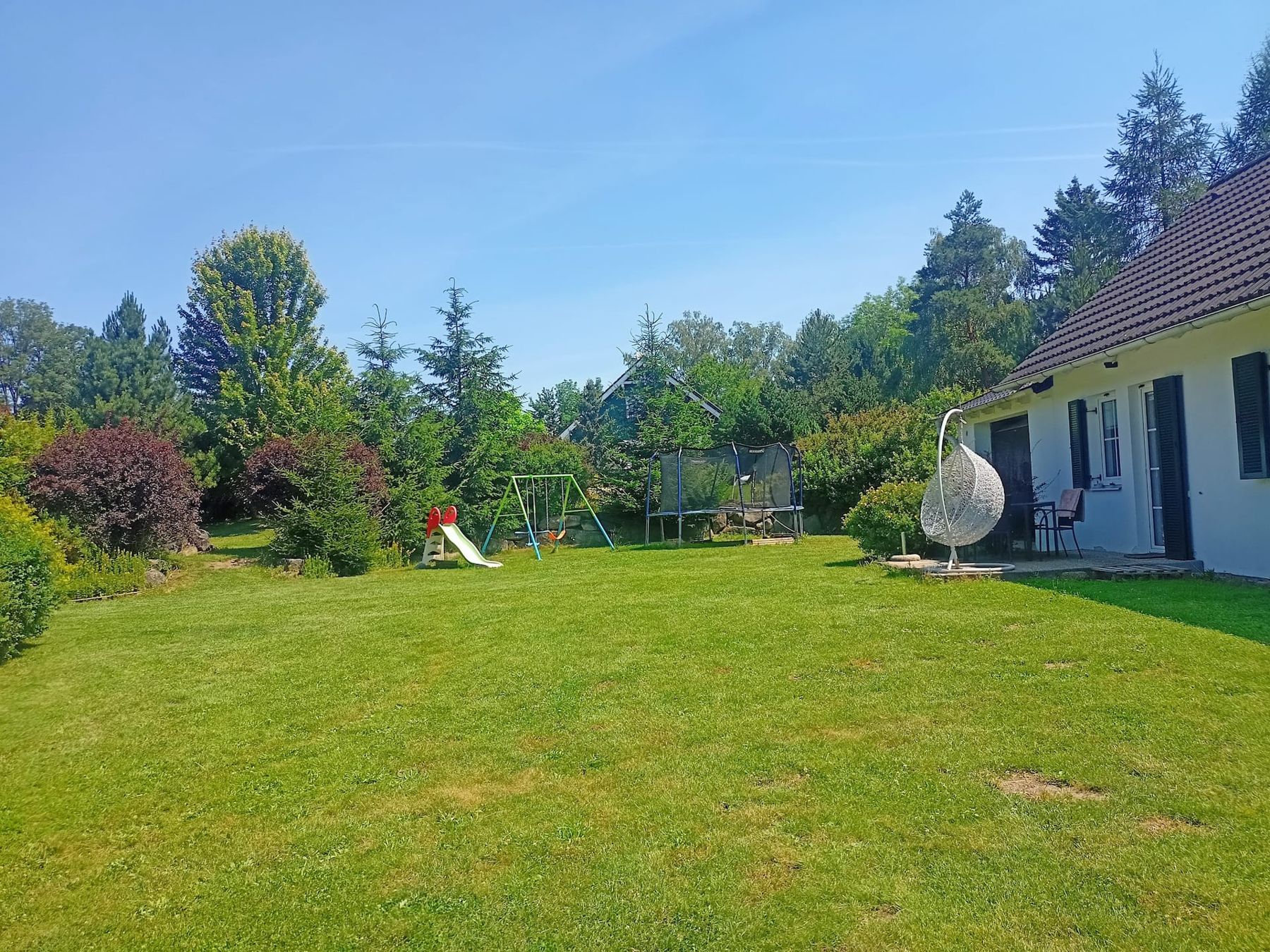 Velký trávník za domem se skluzavkou, houpačkami, trampolínou a závěsným křeslem na terase